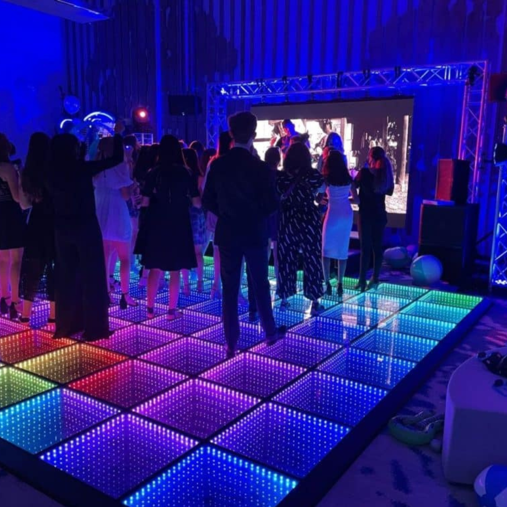 Guests dancing on a vibrant multicolour LED Dance Floor at an indoor event with stage lighting and a large video screen.