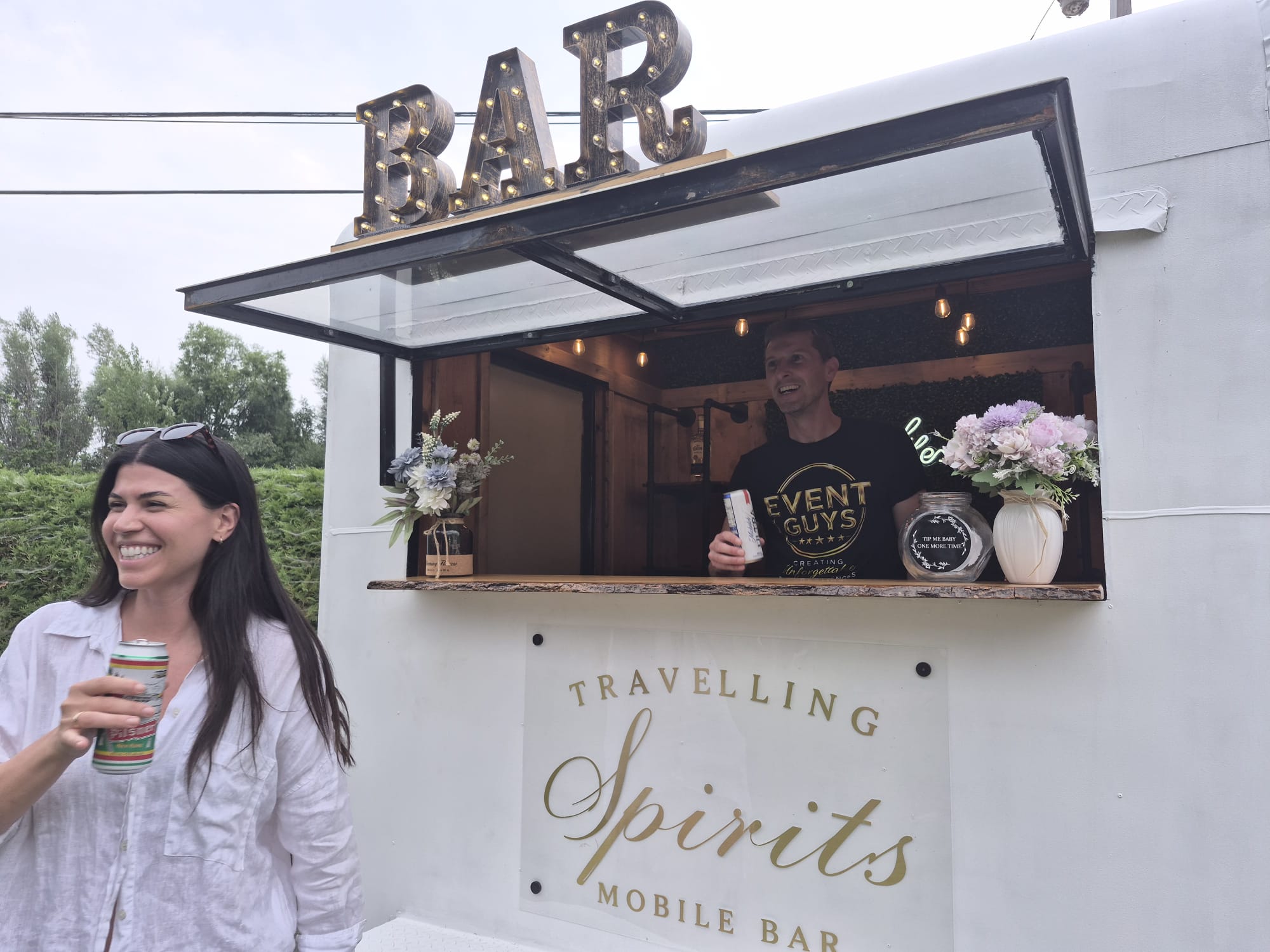 Smiling bartender serving canned drinks to a guest at the Travelling Spirits mobile bar trailer window