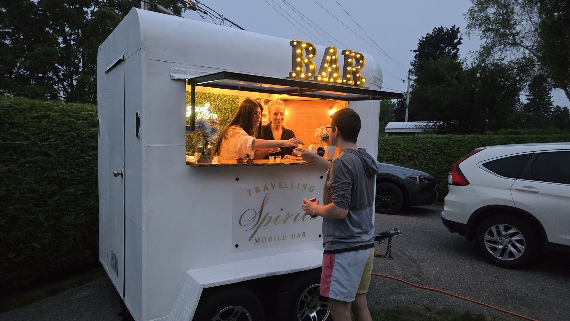 Bartenders serving drinks at dusk from the Travelling Spirits mobile bar trailer service window