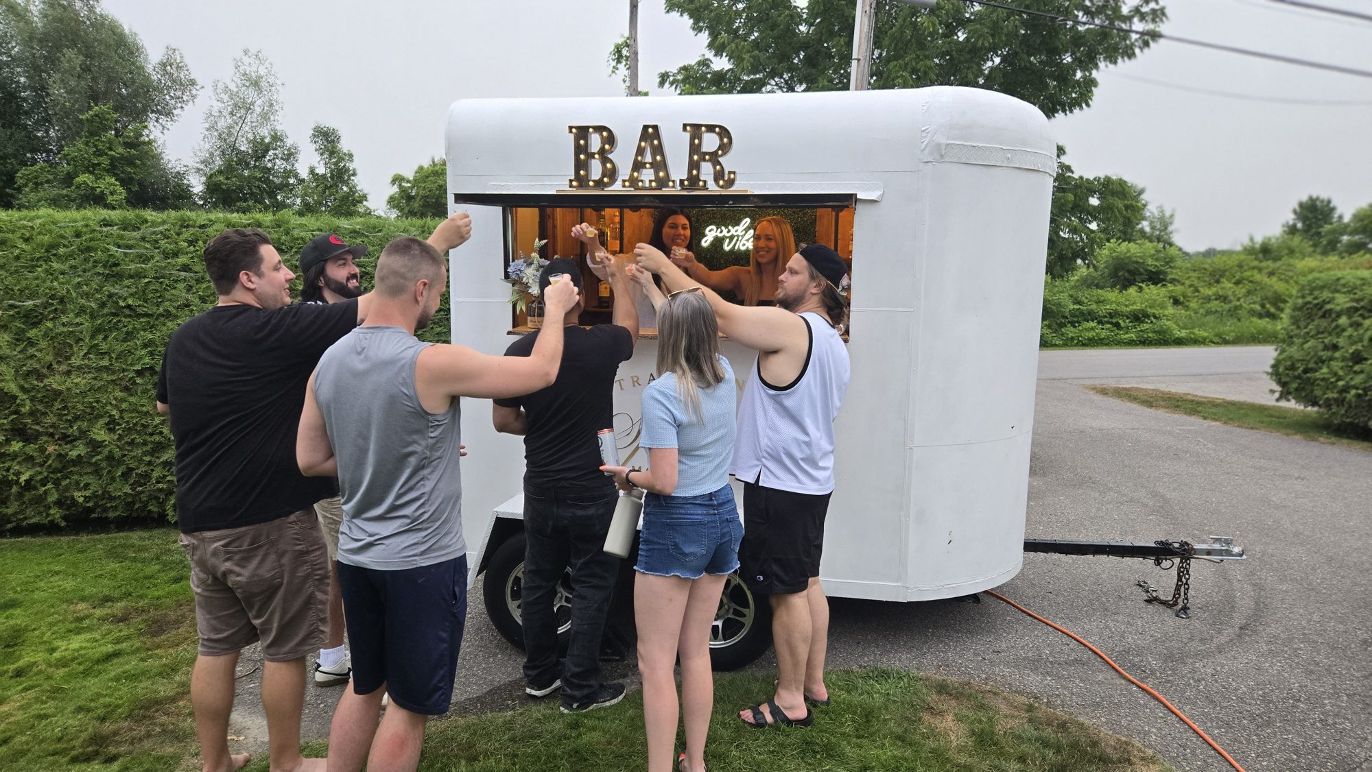 Group of wedding guests raising drinks in a toast at the Travelling Spirits mobile bar trailer service window