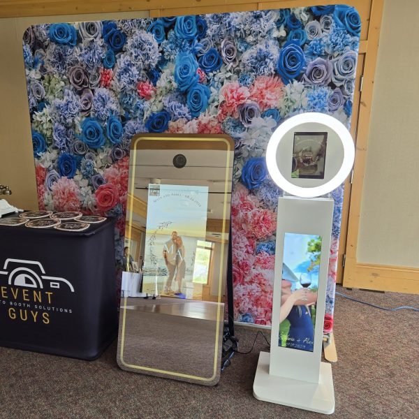 Photo Booth Services for Weddings, Corporate Events, and Parties Photo booth display setup featuring the Event Guys Magic Mirror Photo Booth, iPad Tower Booth, and floral backdrop at an event.